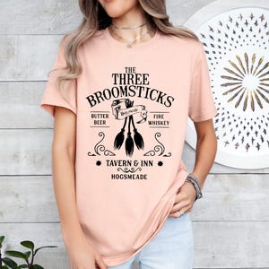May include: A light peach-colored t-shirt featuring a black graphic design. The design includes the text "THE THREE BROOMSTICKS" above an illustration of three brooms. Below the brooms are the words "TAVERN & INN HOGSMEADE".