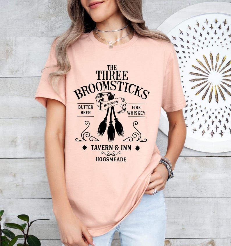 May include: A light peach-colored t-shirt featuring a black graphic design. The design includes the text "THE THREE BROOMSTICKS" above an illustration of three brooms. Below the brooms are the words "TAVERN & INN HOGSMEADE".