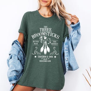 May include: A dark green t-shirt with the text "The Three Broomsticks Tavern & Inn Hogsmeade" in white. The shirt features a graphic of three broomsticks and the words "Butter Beer" and "Fire Whiskey".