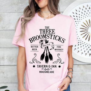 May include: A light pink t-shirt featuring the text "The Three Broomsticks Tavern & Inn Hogsmeade" in black. The design includes three broomsticks and the words "Butter Beer" and "Fire Whiskey".