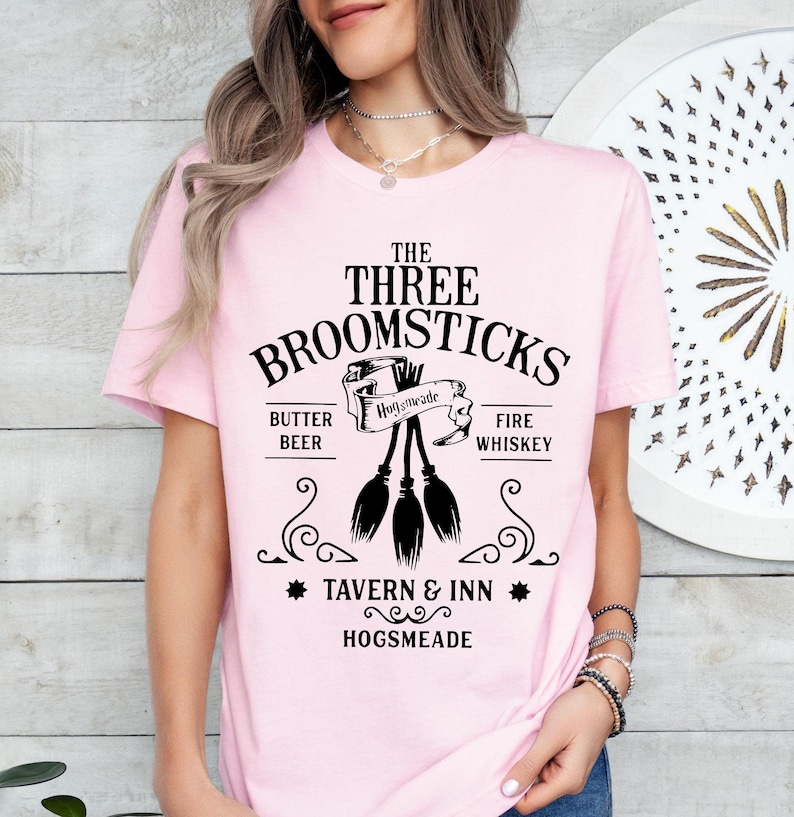 May include: A light pink t-shirt featuring the text "The Three Broomsticks Tavern & Inn Hogsmeade" in black. The design includes three broomsticks and the words "Butter Beer" and "Fire Whiskey".