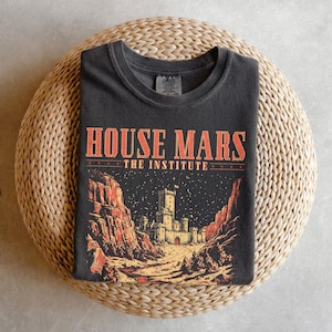 Könnte beinhalten: Ein anthrazitfarbenes T-Shirt mit einem Grafikdruck, der den Text "HOUSE MARS THE INSTITUTE" in Orange zeigt. Das Design stellt eine steinerne Burg zwischen zwei felsigen Klippen unter einem Sternenhimmel in Orange- und Gelbtönen dar.