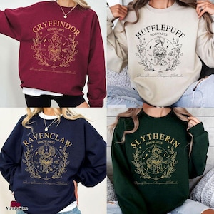 May include: Four sweatshirts, each featuring a different Hogwarts house crest. The sweatshirts are in burgundy, cream, navy blue, and dark green. Each design includes the house name, a crest, and botanical elements in gold.