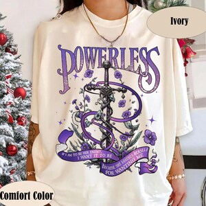 Può includere: Una t-shirt color avorio con una grafica di una spada intrecciata con fiori e nastri viola. Il testo recita POWERLESS, IF I AM TO BE HER ENEMY, I WANT IT TO BE BECAUSE SHE LOATHES HERSELF FOR WANTING ME. La maglietta ha un girocollo.