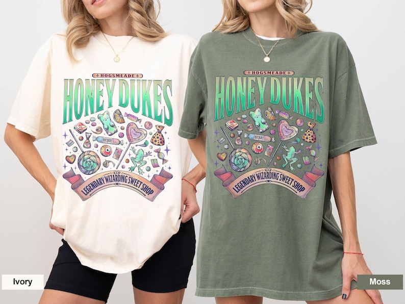May include: Ivory and moss green t-shirts with a graphic design featuring the text "Honeydukes" and various candies. The design includes lollipops, gummy bears, and a banner that reads "Legendary Wizarding Sweet Shop."