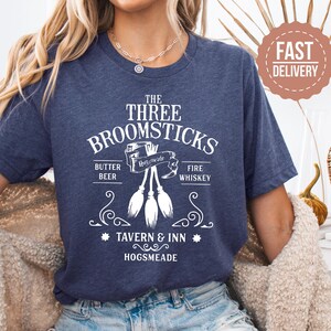 May include: Navy blue t-shirt with a white graphic design. The design features the text "THE THREE BROOMSTICKS" above an illustration of three brooms. Below the brooms are the words "BUTTER BEER" and "FIRE WHISKEY". The text "TAVERN & INN HOGSMEADE" is also included.
