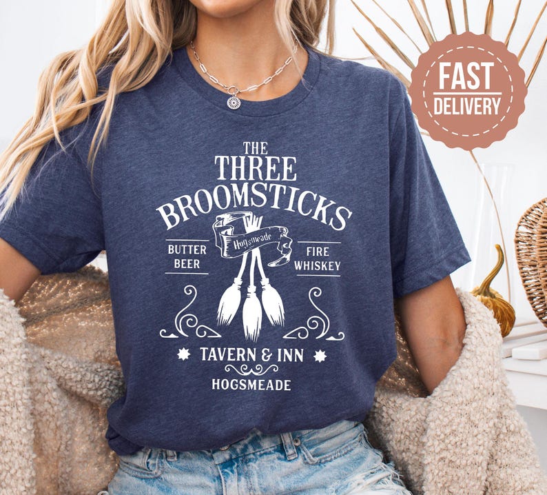 May include: Navy blue t-shirt with a white graphic design. The design features the text "THE THREE BROOMSTICKS" above an illustration of three brooms. Below the brooms are the words "BUTTER BEER" and "FIRE WHISKEY". The text "TAVERN & INN HOGSMEADE" is also included.