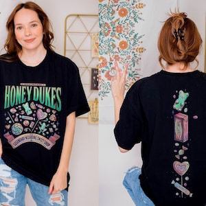May include: Black t-shirt with the text "HONEY DUKES" in green and blue, featuring a colorful illustration of candies and sweets. The back of the shirt has a vertical design with a gummy bear, a box, and various candies.