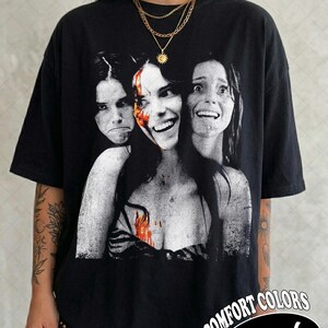May include: A black t-shirt featuring a graphic print of three faces in a monochromatic style with a splash of red color on the central face. The shirt has short sleeves and a crew neckline. The text COMFORT COLORS Black is visible in the corner.