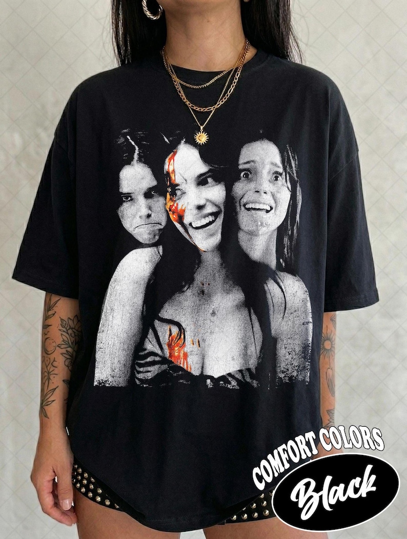 May include: A black t-shirt featuring a graphic print of three faces in a monochromatic style with a splash of red color on the central face. The shirt has short sleeves and a crew neckline. The text COMFORT COLORS Black is visible in the corner.