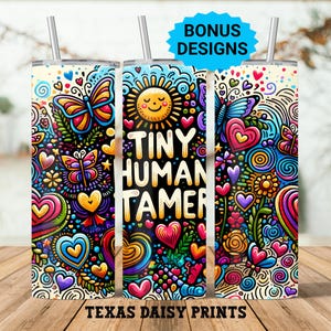 Op de afbeelding: Drie kleurrijke geïsoleerde bekers met deksels en rietjes. De bekers hebben een levendig ontwerp met vlinders, harten, bloemen en de tekst "TINY HUMAN TAMER". De bekers zijn versierd met de woorden "BONUS DESIGNS" en "TEXAS DAISY PRINTS".