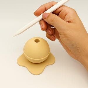 May include: A white Apple Pencil is held over a beige silicone pencil holder shaped like a melting ice cream cone. The holder has a hole in the top for the pencil to be placed in.