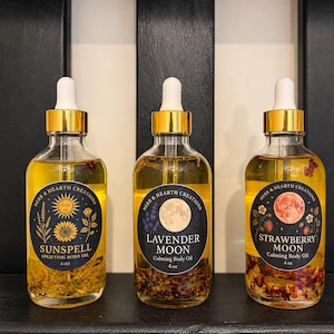 May include: Three clear glass bottles of body oil with gold dropper caps. Each 4 oz bottle features a dark circular label with botanical illustrations and text: Sunspell Uplifting Body Oil, Lavender Moon Calming Body Oil, and Strawberry Moon Calming Body Oil.