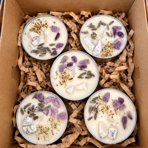 May include: Five round candles in metal tins arranged inside a cardboard box filled with brown paper filler. Each white wax candle is topped with purple amethyst stones, white howlite stones, dried lavender buds, and small dried flower petals.