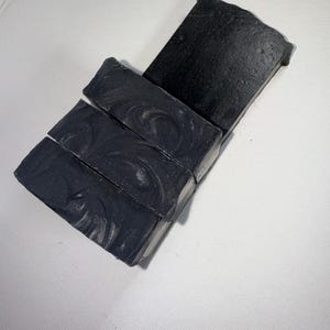 May include: A stack of four rectangular, dark gray soap bars with a marbled design. The bars are arranged diagonally, with the top bar slightly offset. The soap has a matte finish and is set against a white background.