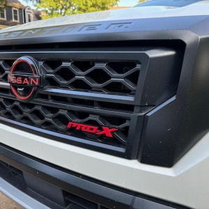 Puede incluir: Primer plano de la parrilla delantera de un coche blanco. La parrilla negra presenta un patrón de panal y un logotipo rojo de Nissan. El texto "PRO-X" es rojo. La palabra "FRONTIER" es visible encima de la parrilla.