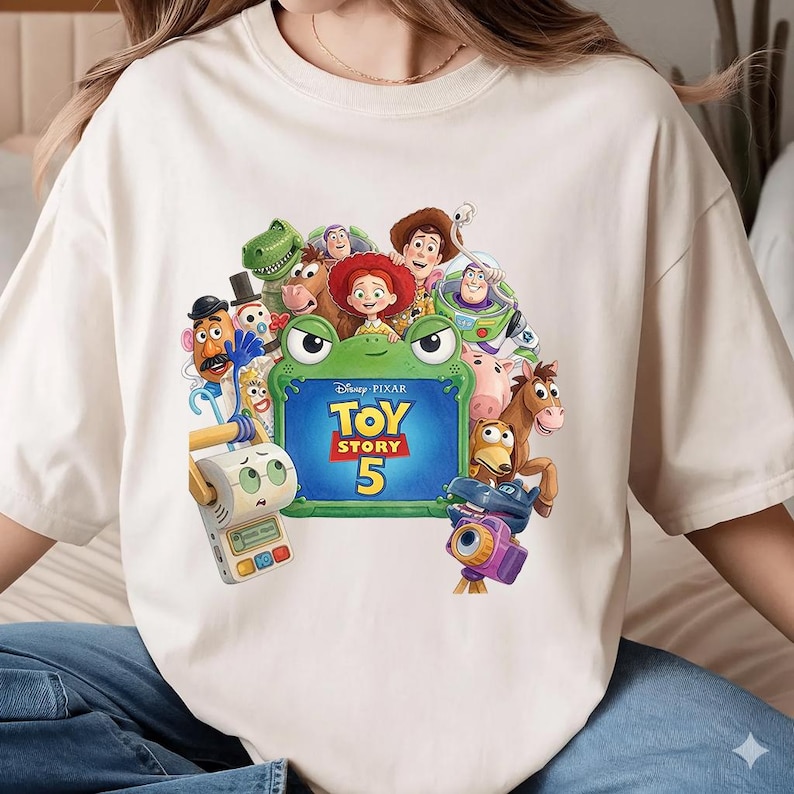 May include: A cream-colored t-shirt featuring a graphic print of various Toy Story characters surrounding a green frog-shaped frame with the text Disney Pixar Toy Story 5. The characters include Woody, Jessie, Buzz Lightyear, Rex, and others.