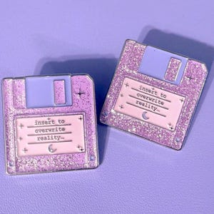 Könnte beinhalten: Zwei glitzernde, lavendelfarbene Disketten-Anstecknadeln mit dem Text "Insert to overwrite reality" in weißer Schrift. Die Anstecknadeln haben einen hellvioletten Schieber und eine kleine Mondgrafik.