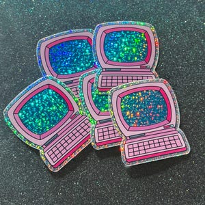 May include: A collection of pink retro computer stickers with holographic screens and keyboards. The stickers have a shiny, iridescent border and are arranged on a glittery black surface. The computer screens display a sparkling, colorful pattern.