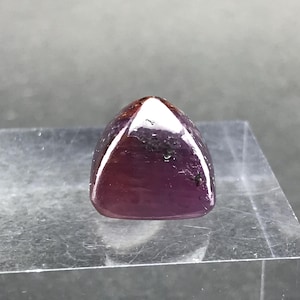 May include: A polished, triangular gemstone in a deep burgundy hue, with a glossy finish. The gem has a rounded top and is set against a clear, angled surface, highlighting its natural beauty. The stone has dark inclusions.