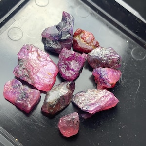 May include: A selection of raw ruby gemstones in shades of pink, red, and purple. The natural stones have a crystalline structure and are displayed on a dark surface. The gemstones vary in size and shape, showcasing their natural beauty.