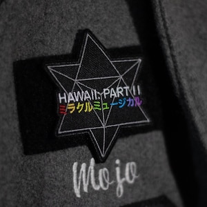 Puede incluir: Un parche negro en forma de estrella con el texto "HAWAII: PART II" y "ミラクルミュージックカル" en colores arcoíris. El parche está cosido en una tela gris con la palabra "Mojo" bordada debajo.