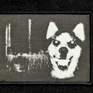 Op de afbeelding: Een zwart-wit patch met een lachende hond met heldere ogen. De rechthoekige patch heeft een zwarte rand en een getextureerd oppervlak. Het gezicht van de hond is het centrale beeld, met een brede grijns en gedetailleerde vacht.