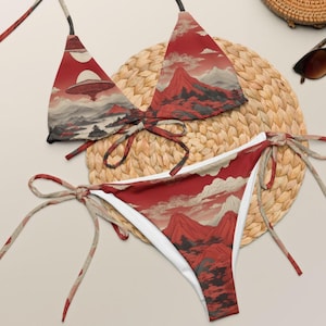 May include: A red and white bikini set with a mountain and cloud landscape design. The top features a UFO graphic. The bikini is tied with string ties. The set is displayed on a woven mat.