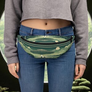 May include: Green fanny pack featuring a repeating pattern of UFOs, trees, and clouds. The bag has a black zip and strap. The background is a light green and white colour scheme.