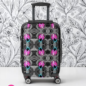 May include: A black rolling suitcase with a repeating pattern of pink and blue ladybugs on a gray background. The suitcase has a retractable handle and four wheels. The background is a black and white floral design.