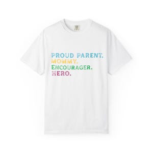 May include: White t-shirt with the text "PROUD PARENT. MOMMY. ENCOURAGER. HERO." in blue, yellow, green, and purple. The shirt has a crew neck and short sleeves. The text is centered on the front of the shirt.