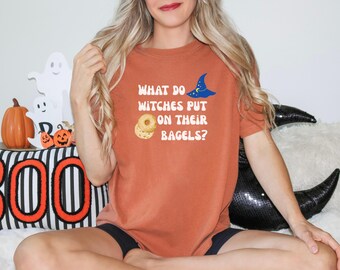 May include: Rust-colored t-shirt with the text "WHAT DO WITCHES PUT ON THEIR BAGELS?" in white, along with a blue witch hat and bagel graphic. Halloween-themed shirt with a playful design.