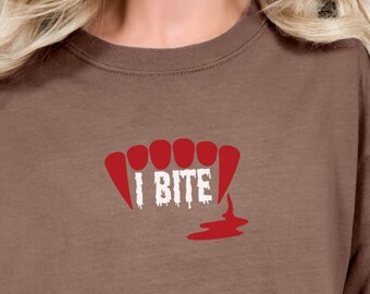 May include: A brown t-shirt featuring a red graphic of vampire fangs with the text "I BITE" in white. Below the fangs is a red blood drop design. The shirt is made of a soft material.