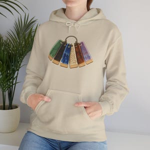 May include: A beige hooded sweatshirt featuring a graphic print of six rectangular ceramic glaze test tiles in green, black, blue, speckled cream, brown, and purple, all attached to a circular metal ring centered on the chest area.