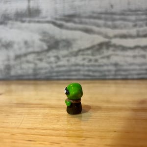 Puede incluir: Peque&ntilde;a figura artesanal de un extraterrestre verde con ojos negros y cuerpo marr&oacute;n. La figura est&aacute; sobre una superficie de madera. El dise&ntilde;o es detallado y probablemente hecho de arcilla o resina.