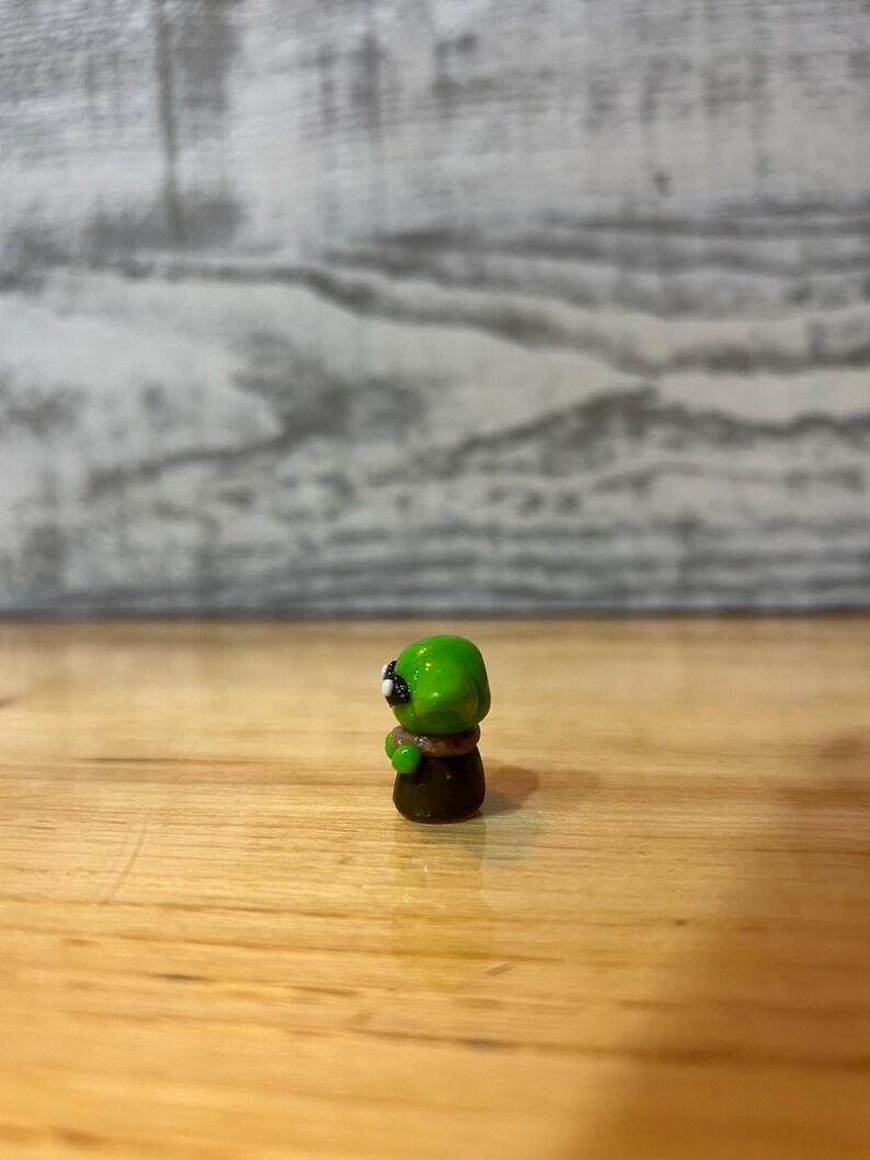 Puede incluir: Peque&ntilde;a figura artesanal de un extraterrestre verde con ojos negros y cuerpo marr&oacute;n. La figura est&aacute; sobre una superficie de madera. El dise&ntilde;o es detallado y probablemente hecho de arcilla o resina.