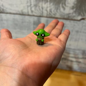 Puede incluir: Una peque&ntilde;a figura verde de un personaje con orejas grandes y ojos negros, con un atuendo marr&oacute;n. La figura se sostiene en una mano, mostrando su tama&ntilde;o y detalle. Es una artesan&iacute;a hecha a mano.