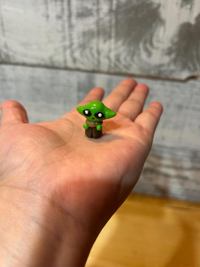 Puede incluir: Una peque&ntilde;a figura verde de un personaje con orejas grandes y ojos negros, con un atuendo marr&oacute;n. La figura se sostiene en una mano, mostrando su tama&ntilde;o y detalle. Es una artesan&iacute;a hecha a mano.