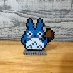 May include: A blue and white pixel art figure of a cartoon character with a brown accessory, standing on a gray base. The character has black outlines, pink cheeks, and a fluffy appearance. The figure is placed on a wooden surface.