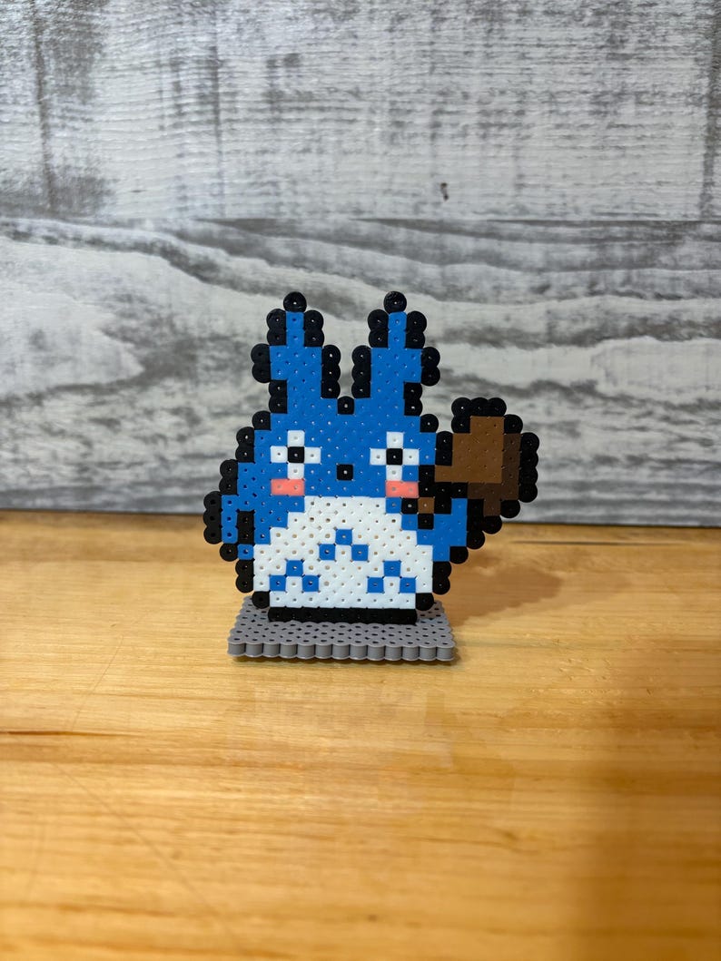 May include: A blue and white pixel art figure of a cartoon character with a brown accessory, standing on a gray base. The character has black outlines, pink cheeks, and a fluffy appearance. The figure is placed on a wooden surface.