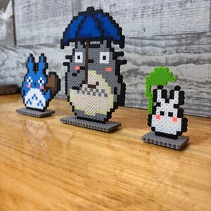 May include: Three pixel art figures from the animated film, "My Neighbor Totoro." The central figure is Totoro, holding a blue umbrella. A blue cat-like creature is on the left, and a white rabbit-like creature is on the right. All are on gray bases.