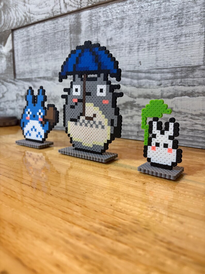 May include: Three pixel art figures from the animated film, "My Neighbor Totoro." The central figure is Totoro, holding a blue umbrella. A blue cat-like creature is on the left, and a white rabbit-like creature is on the right. All are on gray bases.