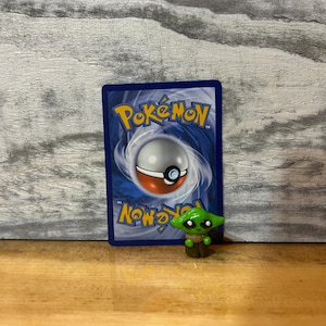 Puede incluir: Una peque&ntilde;a figura verde de un personaje con ojos grandes y orejas puntiagudas, frente a una carta Pok&eacute;mon azul. La carta tiene un dise&ntilde;o de Pokeball blanco y rojo y texto amarillo.