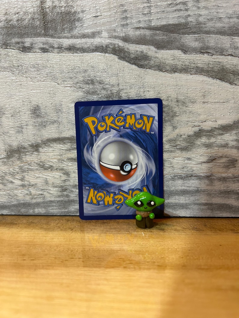 Puede incluir: Una peque&ntilde;a figura verde de un personaje con ojos grandes y orejas puntiagudas, frente a una carta Pok&eacute;mon azul. La carta tiene un dise&ntilde;o de Pokeball blanco y rojo y texto amarillo.
