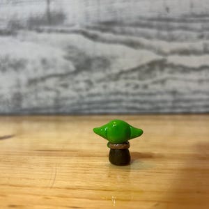 Puede incluir: Peque&ntilde;a figura hecha a mano de un personaje con orejas grandes y cabeza verde. La figura est&aacute; sobre una superficie de madera. El personaje tiene un cuerpo marr&oacute;n y una cabeza verde con orejas grandes.