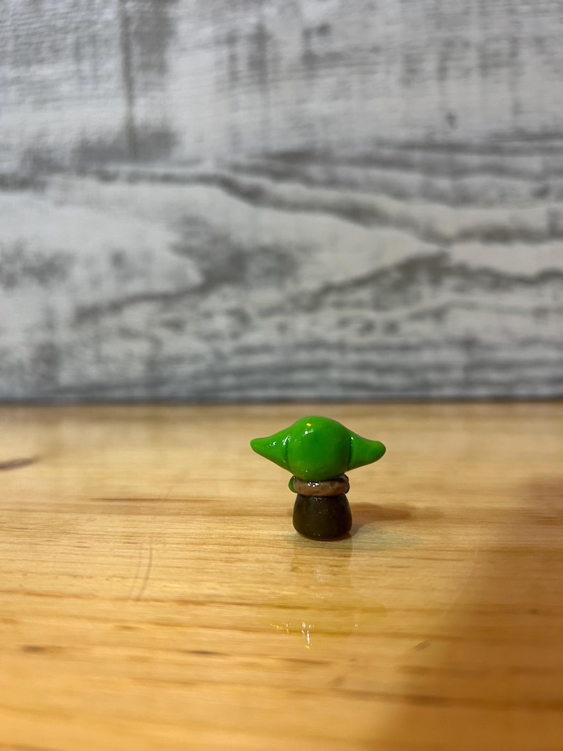 Puede incluir: Peque&ntilde;a figura hecha a mano de un personaje con orejas grandes y cabeza verde. La figura est&aacute; sobre una superficie de madera. El personaje tiene un cuerpo marr&oacute;n y una cabeza verde con orejas grandes.