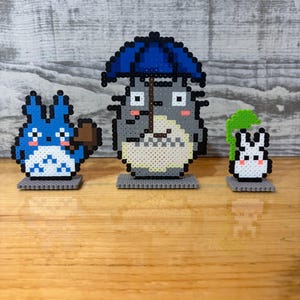 May include: Three pixel art figures from the animated film, My Neighbor Totoro, are displayed on a wooden surface. The central figure is Totoro, holding a blue umbrella. A blue creature with a broom and a small white rabbit are also present.