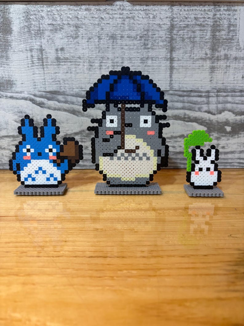 May include: Three pixel art figures from the animated film, My Neighbor Totoro, are displayed on a wooden surface. The central figure is Totoro, holding a blue umbrella. A blue creature with a broom and a small white rabbit are also present.