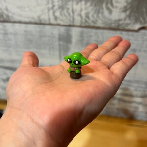 Puede incluir: Una peque&ntilde;a figura verde de un personaje con orejas grandes y ojos negros, sostenida en una mano abierta. La figura tiene una base marr&oacute;n y una prenda de color beige. La figura es una representaci&oacute;n en miniatura de un personaje popular de una franquicia de ciencia ficci&oacute;n.