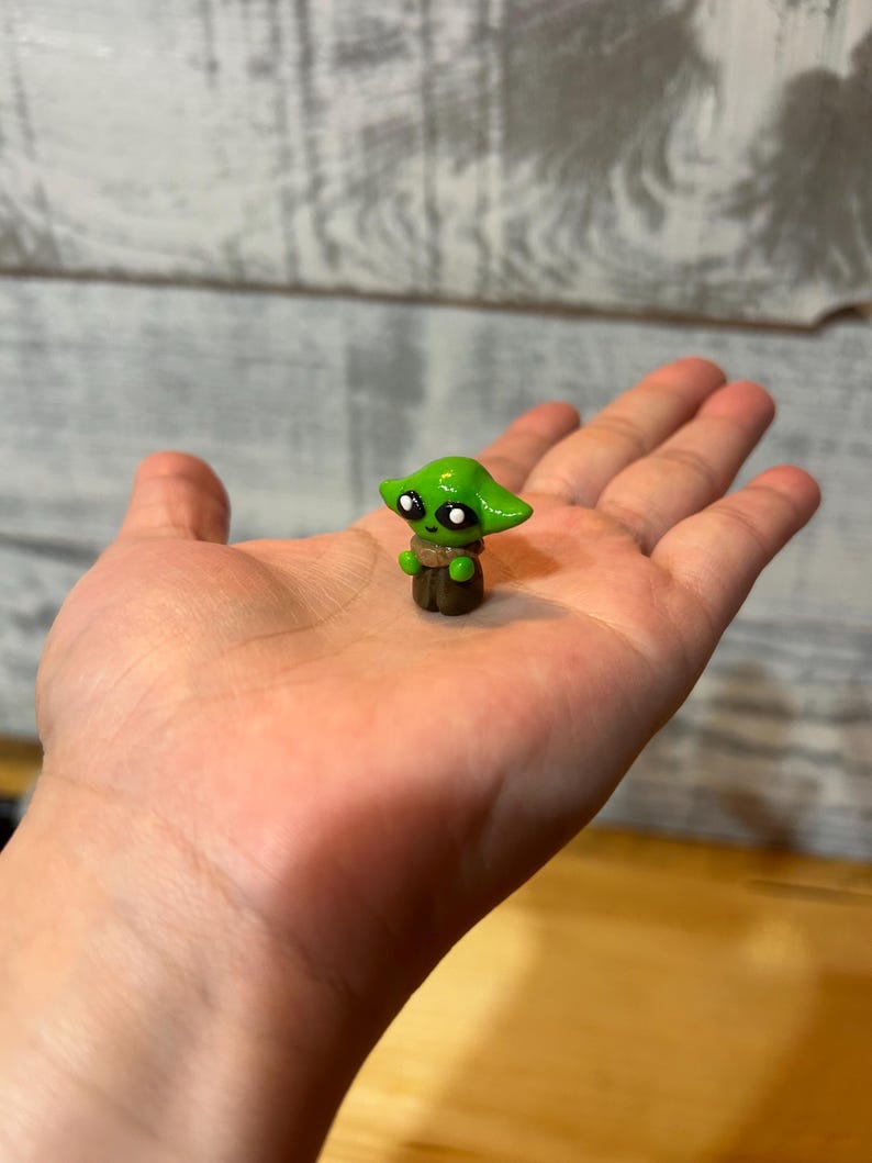 Puede incluir: Una peque&ntilde;a figura verde de un personaje con orejas grandes y ojos negros, sostenida en una mano abierta. La figura tiene una base marr&oacute;n y una prenda de color beige. La figura es una representaci&oacute;n en miniatura de un personaje popular de una franquicia de ciencia ficci&oacute;n.